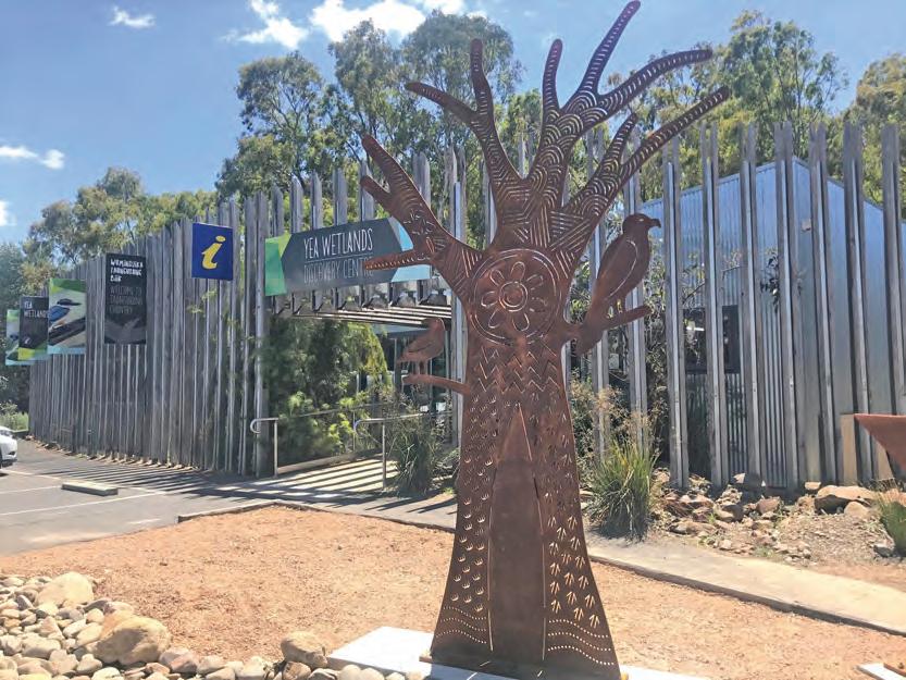 metal sculpture of a tree with indigenous markings on it out the front of a modern looking building with leafy trees in the background. The ground that the sculpture is on is light brown and sandy, like the desert.