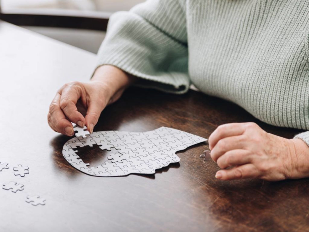 hands of elderly woman shown putting a jigsaw puzzle together that is in the shape of a head, on a brown wooden table.
